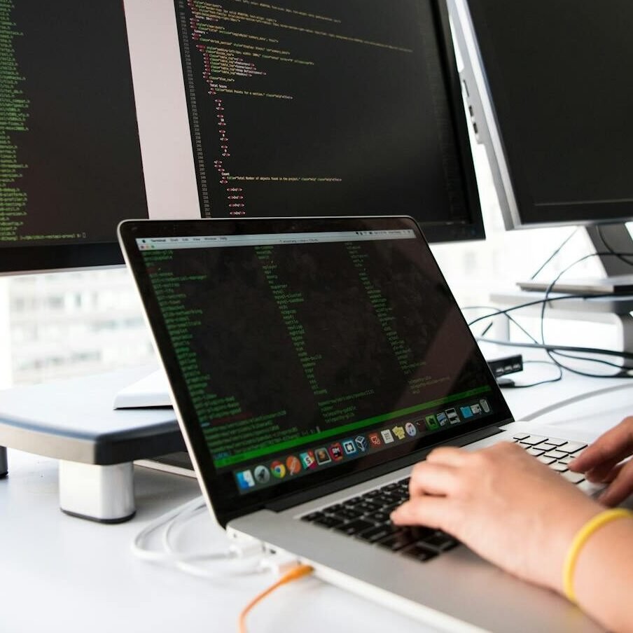 A developer writes code on a laptop in front of multiple monitors in an office setting.